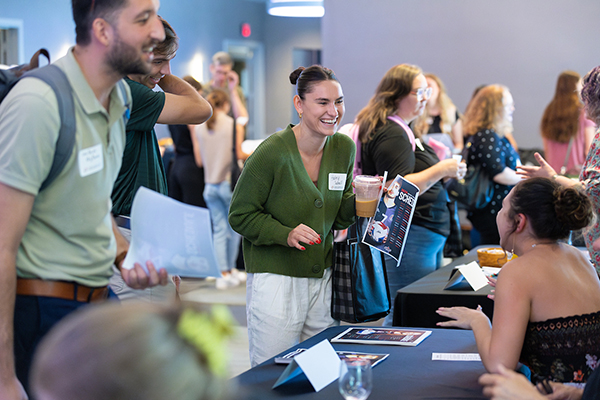 a woman in a green cardigan and a man in a green shirt wearing a backpack have a friendly chat at a new student event