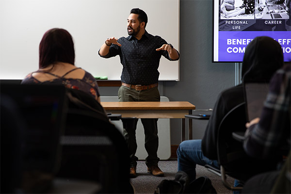 teacher gesturing while standing at the front of a classroom as students look on
