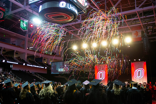 dark indoor arena with confetti streamers shooting over the heads of UNO graduates in cap and gowns