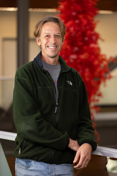 Smiling man in a dark green fleece jacket leaning on a railing indoors, with a blurred red sculpture in the background.