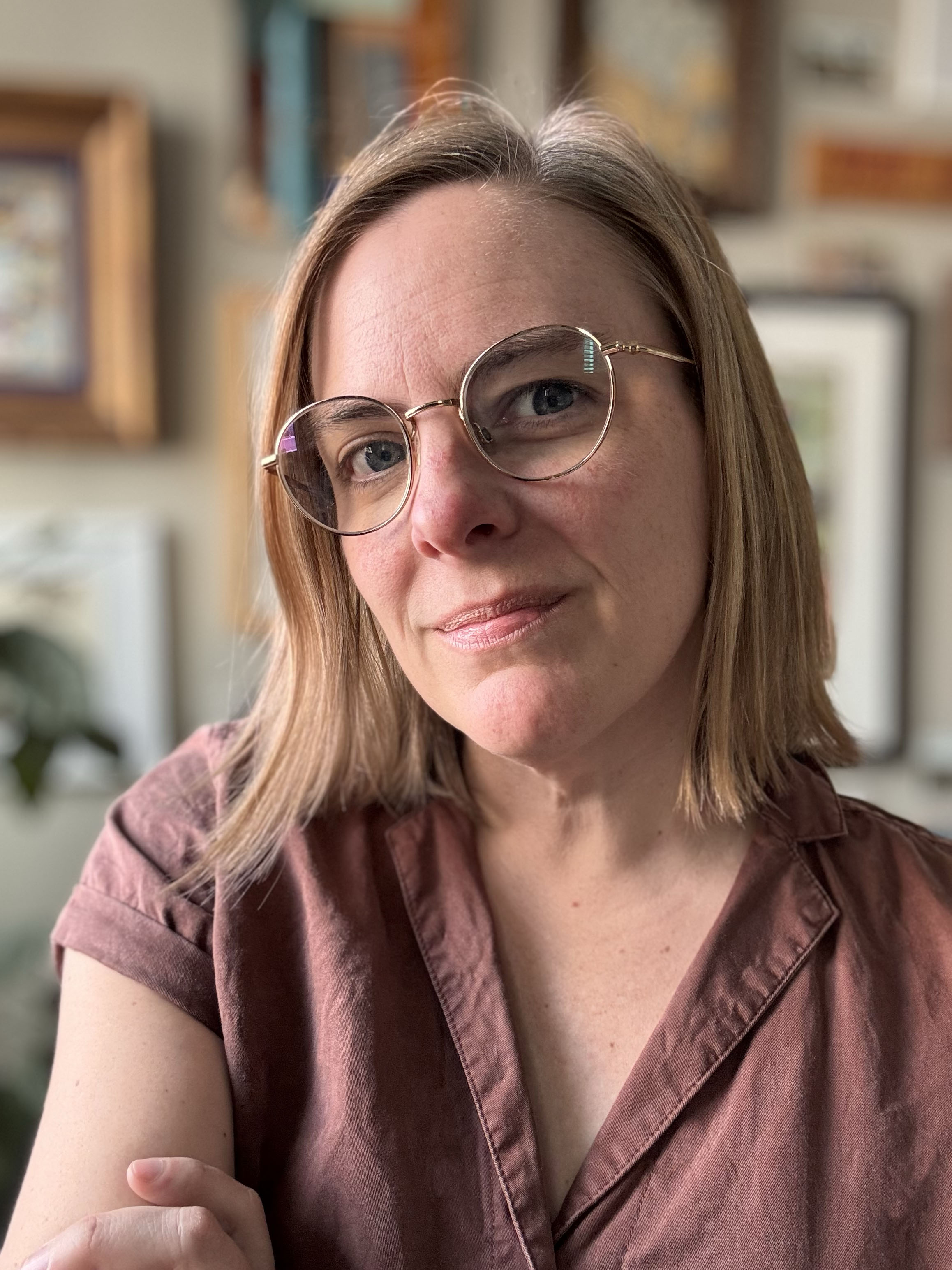 Close-up portrait of a woman with shoulder-length blonde hair wearing round glasses and a brown collared shirt, with a softly blurred indoor background.