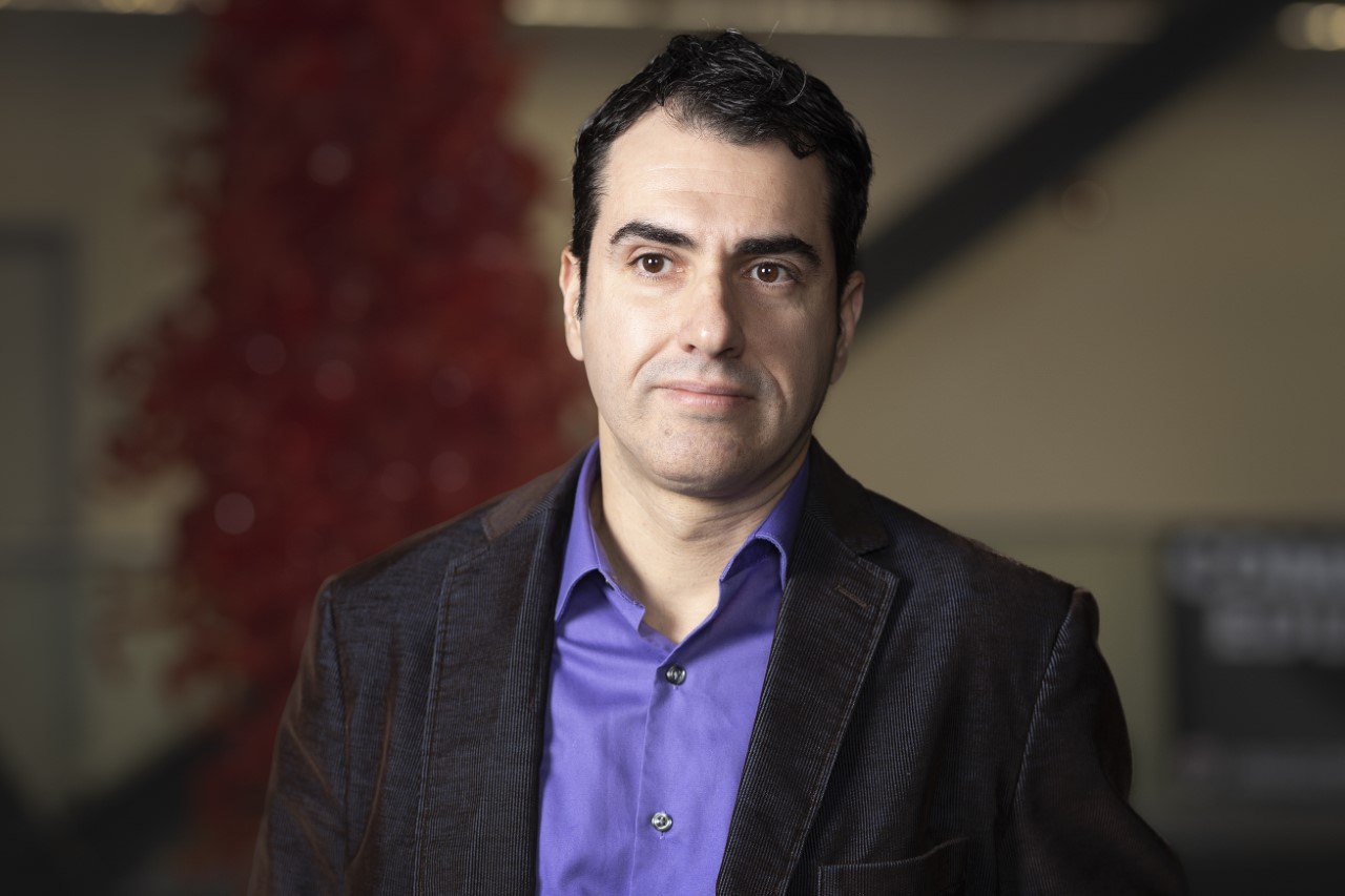 A man with dark hair and a purple shirt looks at the camera