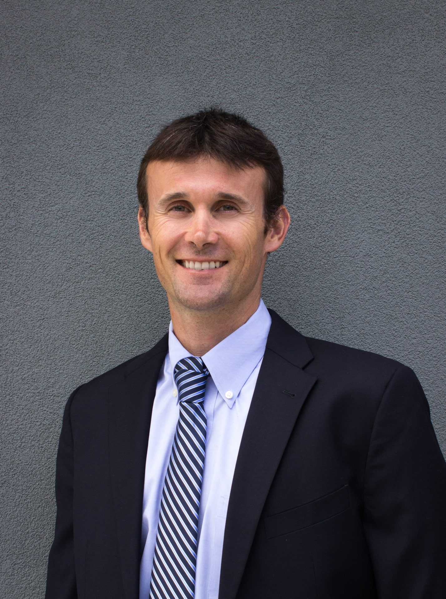 A man with dark hair wearing a suit and tie looks at the camera