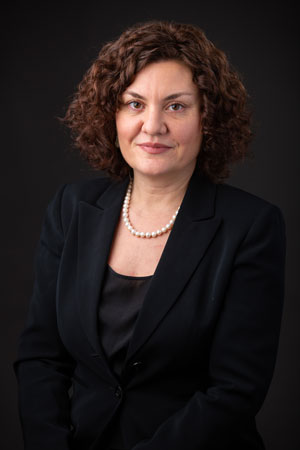 Studio portrait of a woman with curly shoulder-length hair wearing a dark blazer and pearl necklace against a black background.