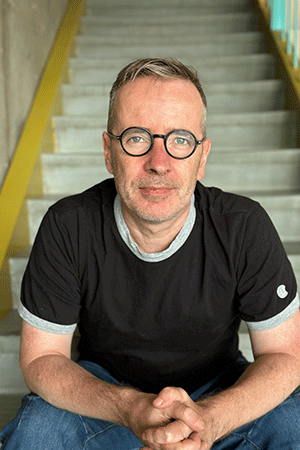 a photo of a man with glasses sitting on a staircase and looking at the camera