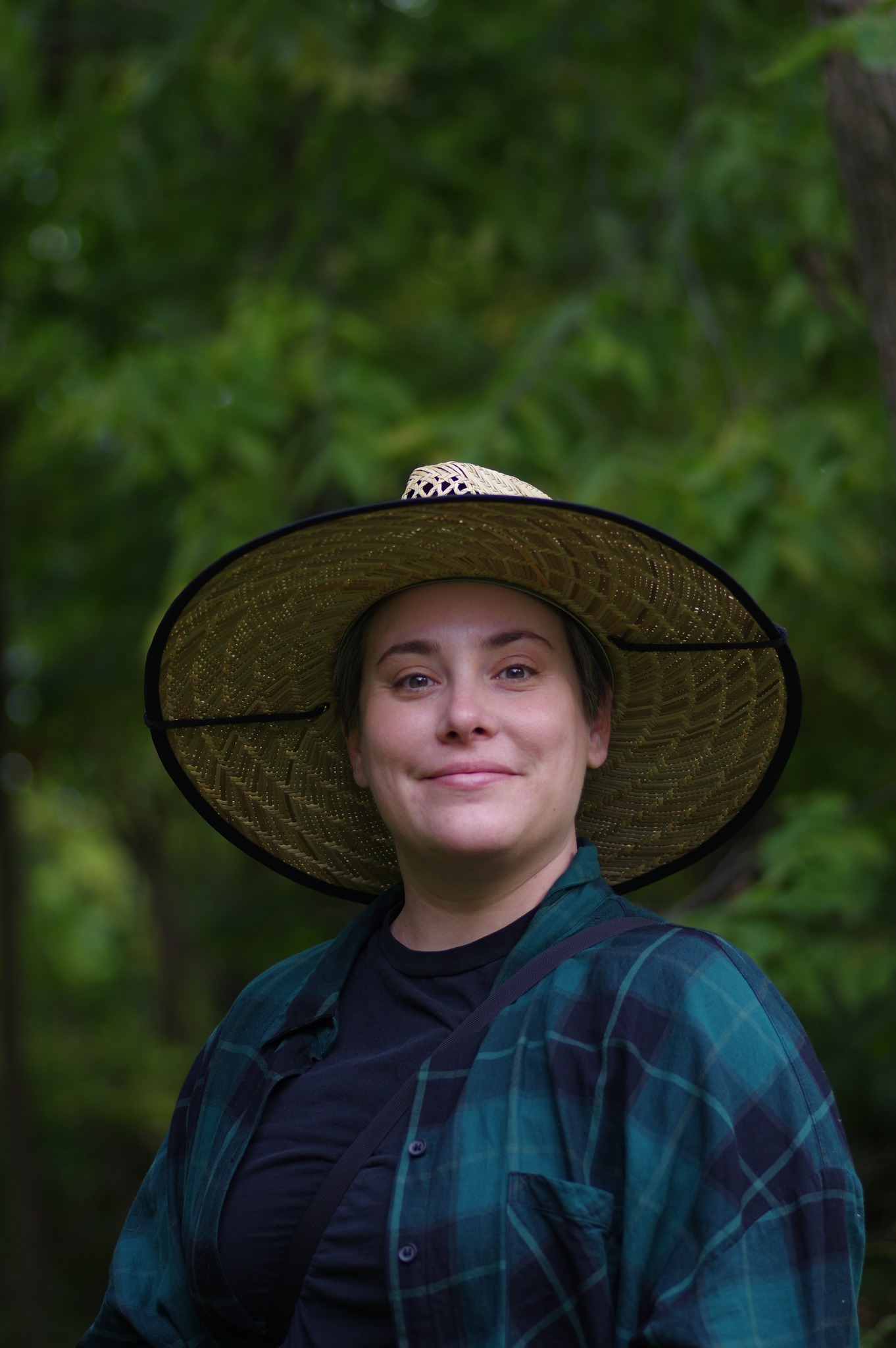 Smiling person wearing a wide-brim straw hat and a green plaid shirt, standing outdoors with blurred green foliage in the background.