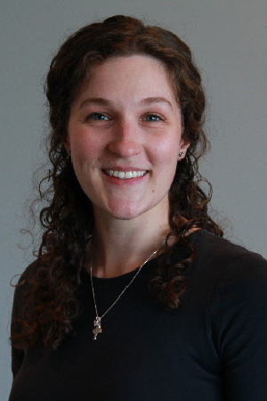 Smiling woman with curly brown hair wearing a dark top and a small cross necklace against a plain gray background.