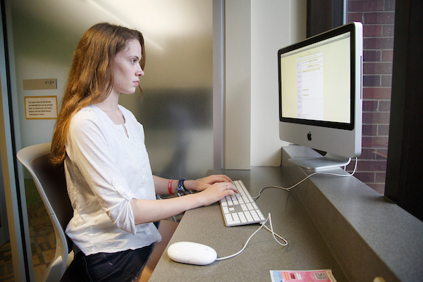 a young woman works on a computer at a desk in front of a bright window.