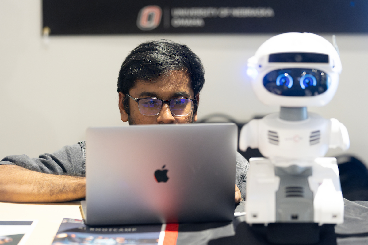 a student works on a laptop next to a small robot.