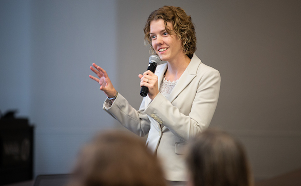 A woman in a light-colored blazer speaks confidently into a microphone, gesturing with her hand while addressing an audience.