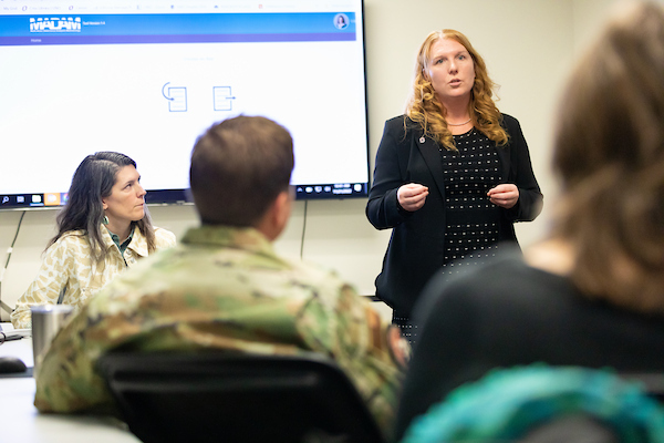 A woman in business attire gives a presentation to an audience that includes individuals in military uniforms. She stands in front of a large screen displaying her presentation, suggesting a professional setting focused on security studies or national defense.