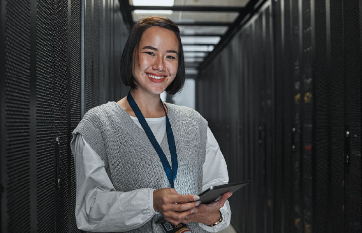Woman holding a tablet and smiling while standing in a server room corridor lined with racks of equipment.