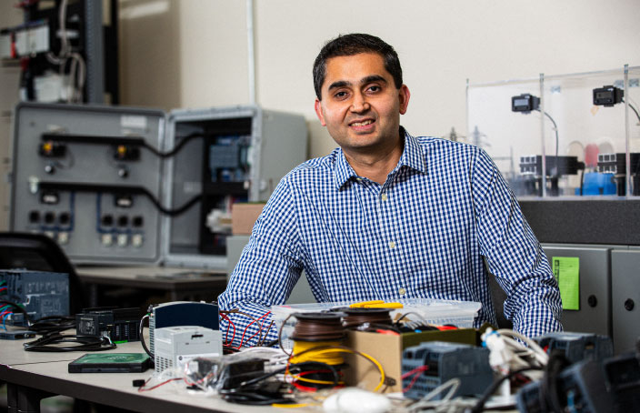 A professor or professional sitting at a workstation with electronic equipment, smiling at the camera in a university lab setting.