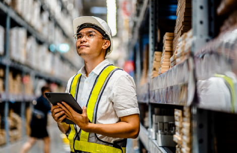 A man in a safety vest and hard hat stands in a warehouse aisle holding a tablet, symbolizing leadership and learning applied in the workplace.