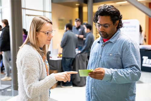 A middle aged woman points to a business card held by a younger man at a career fair