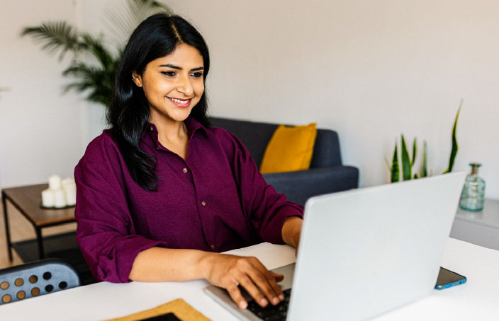 A woman in a burgundy blouse smiles while working on a laptop at a table in a modern, well-lit living space, representing flexible online learning.