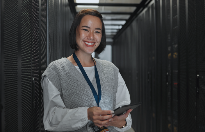 Smiling cybersecurity professional in a data center, holding a tablet and wearing an access badge.