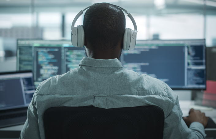 A person wearing headphones sits at a desk, focused on computer screens displaying lines of code