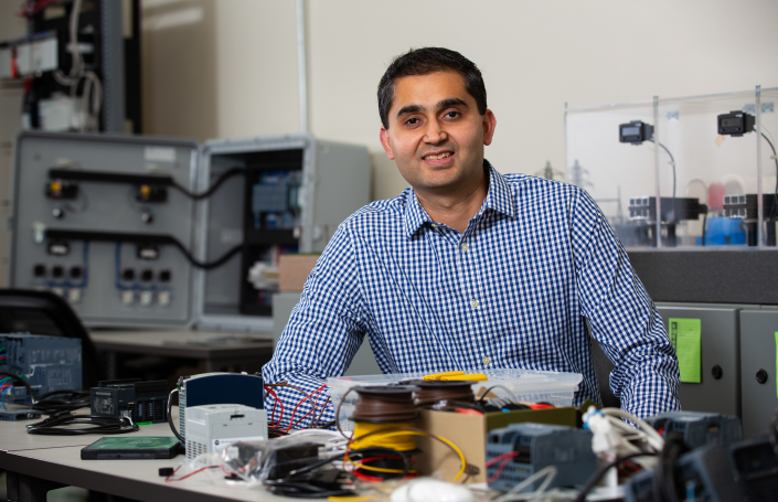 A professor or professional sitting at a workstation with electronic equipment, smiling at the camera in a university lab setting.