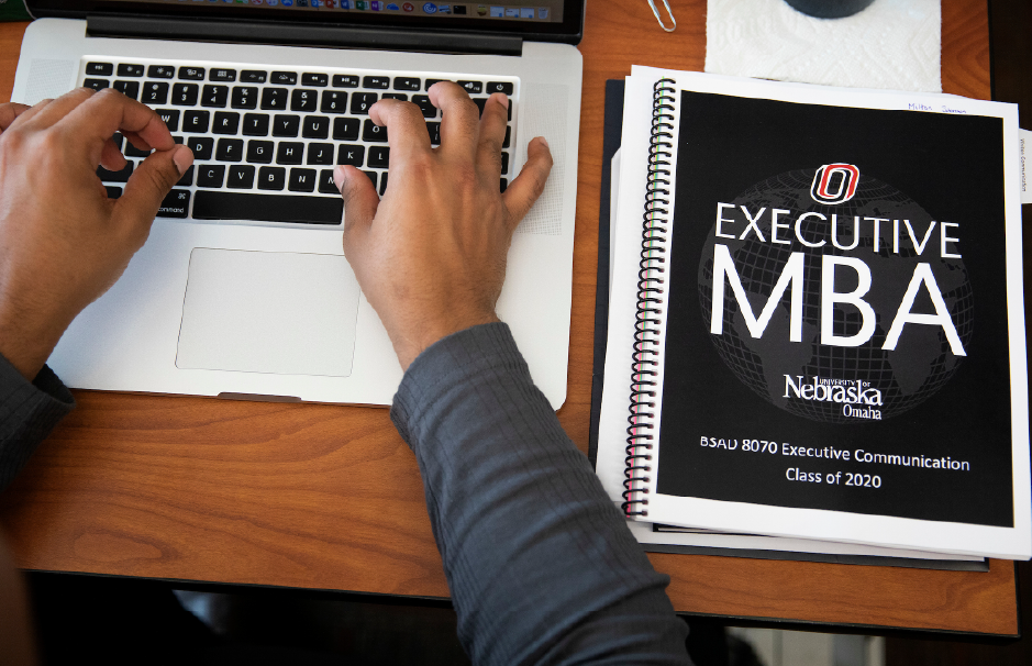 A close-up of hands typing on a laptop next to a spiral-bound Executive MBA coursebook from the University of Nebraska Omaha.