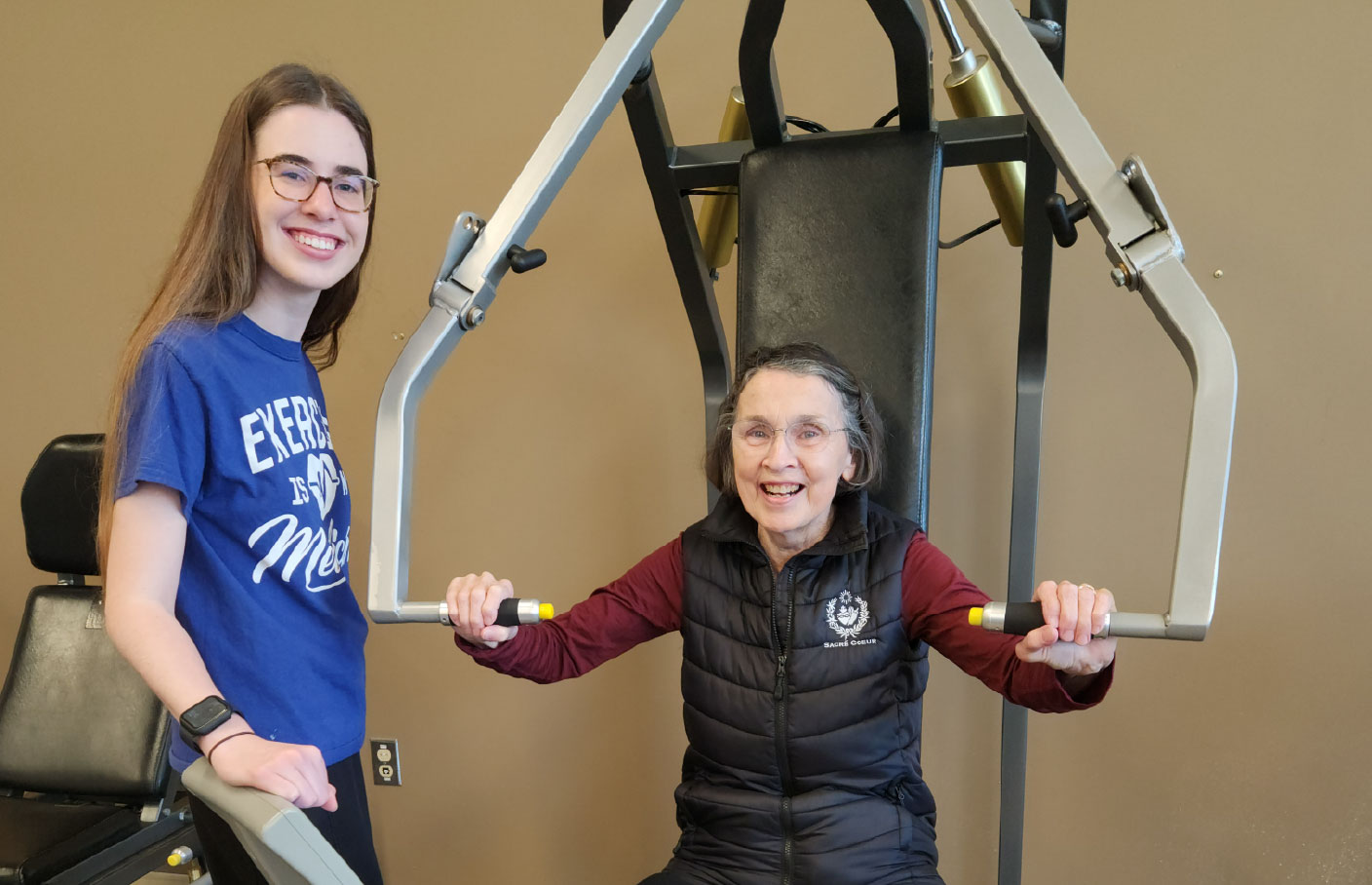 A young woman assists an older woman using weight-lifting equipment, both smiling and enjoying a workout session.