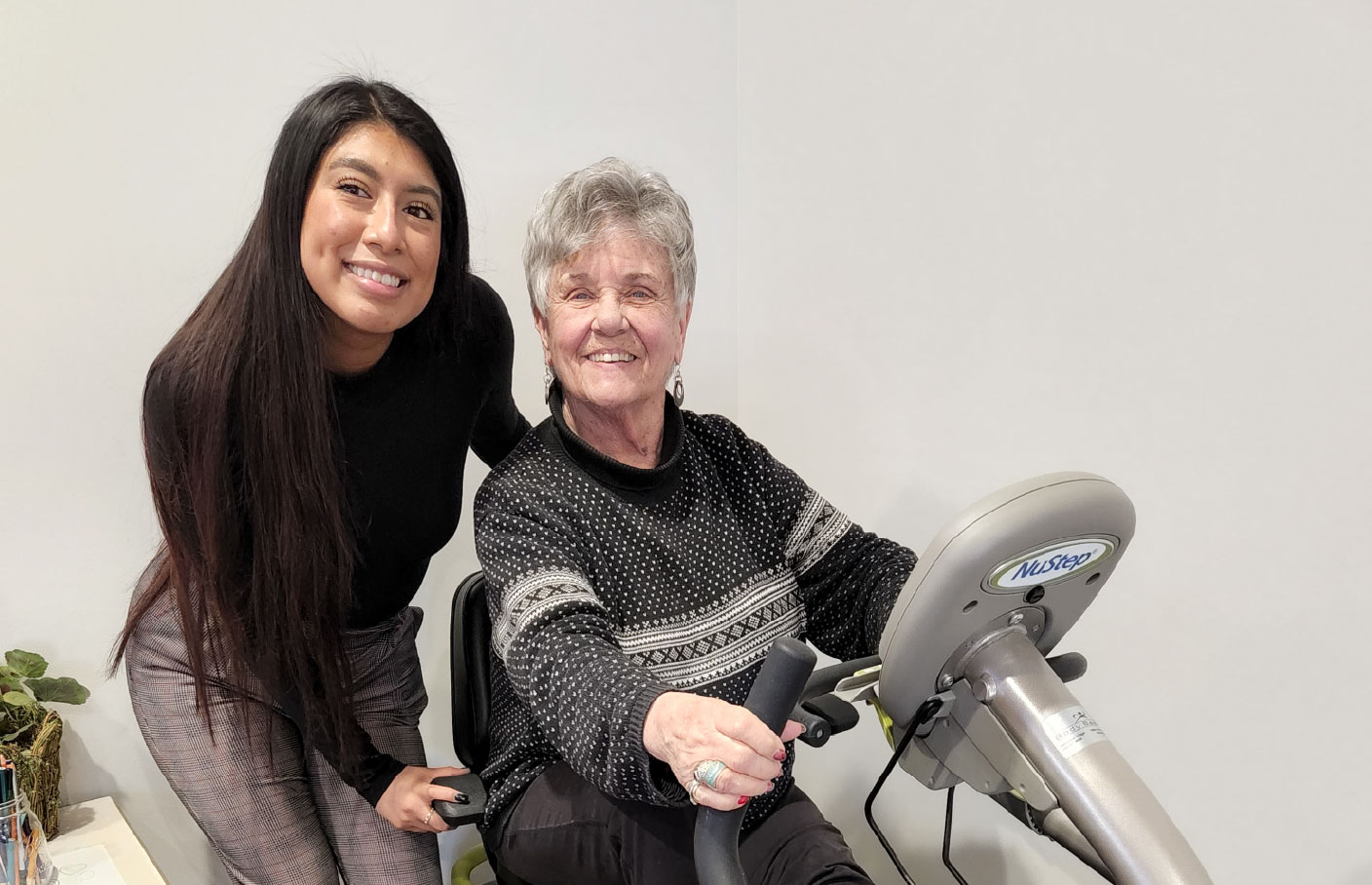 A young woman assists an older woman using weight-lifting equipment, both smiling and enjoying a workout session.