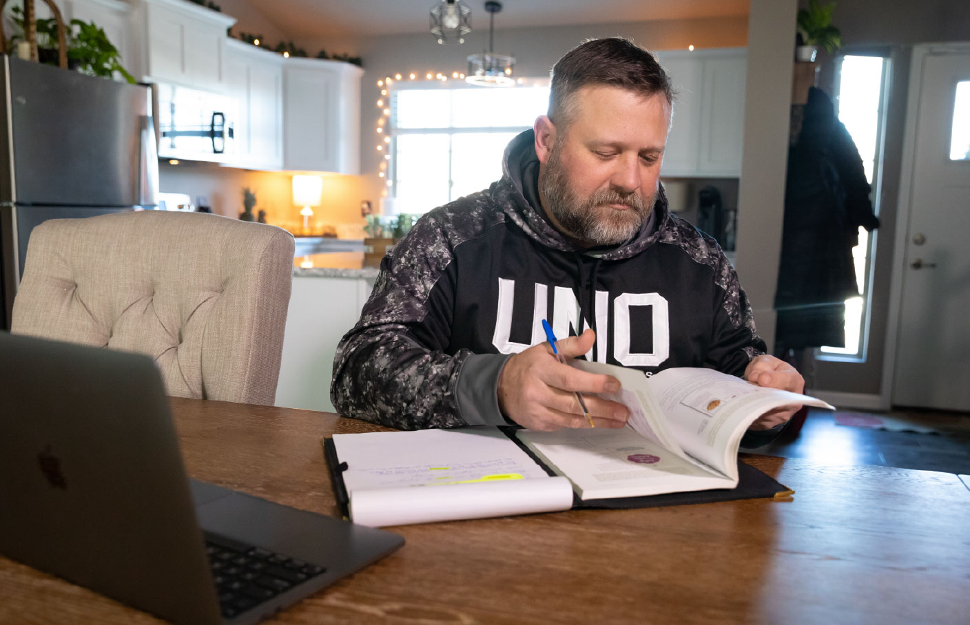 A man wearing a University of Nebraska Omaha hoodie studies at a kitchen table, reviewing a textbook and taking notes, with a laptop open beside him in a home setting.