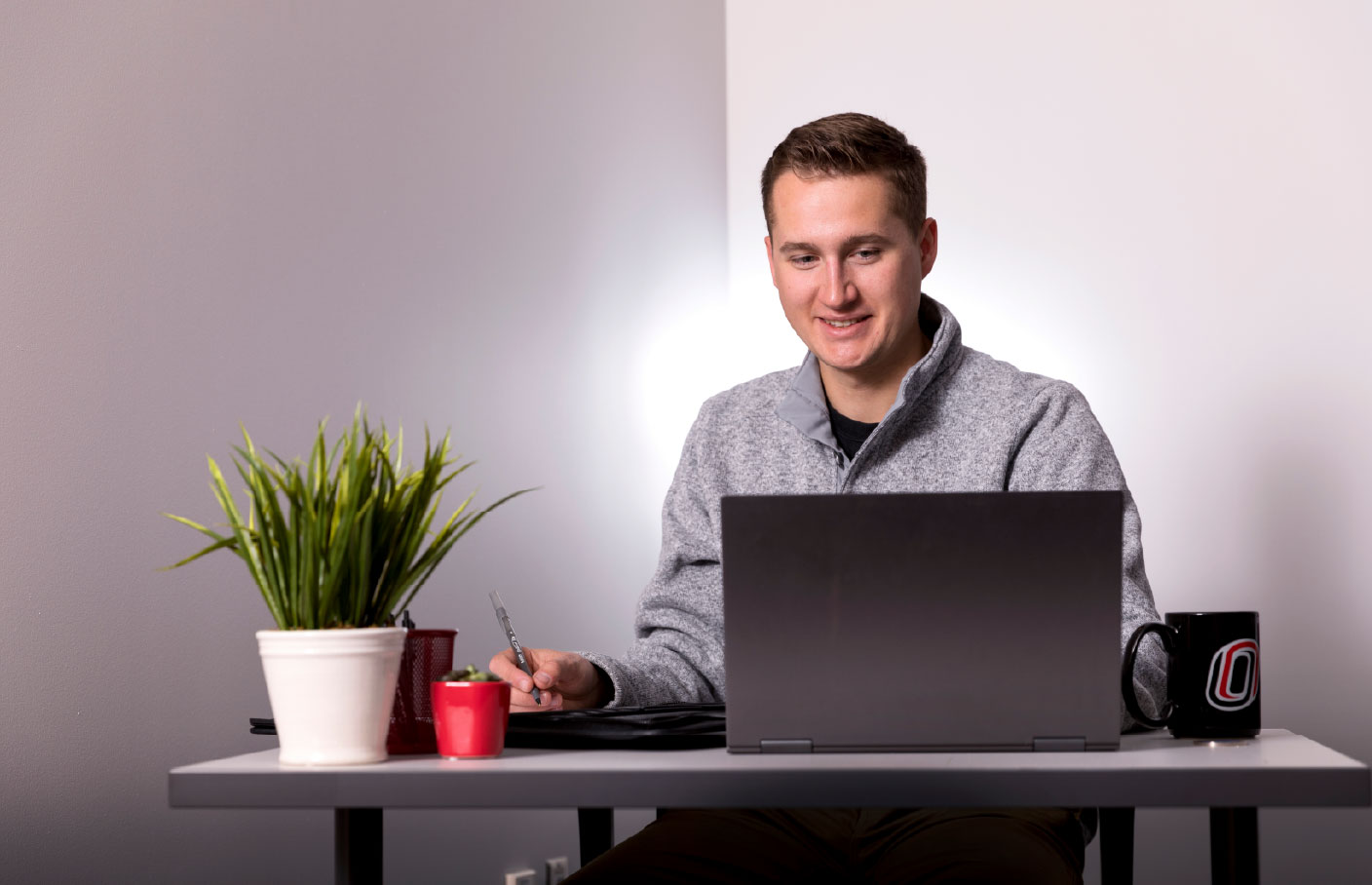 A man smiles while working at a desk with a laptop, accompanied by a potted plant and a University of Nebraska Omaha mug.