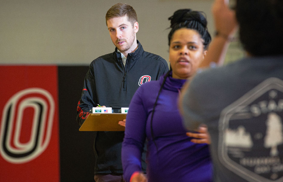 A male instructor in a UNO-branded jacket observes students in a gym setting, clipboard in hand, as a fitness activity takes place.