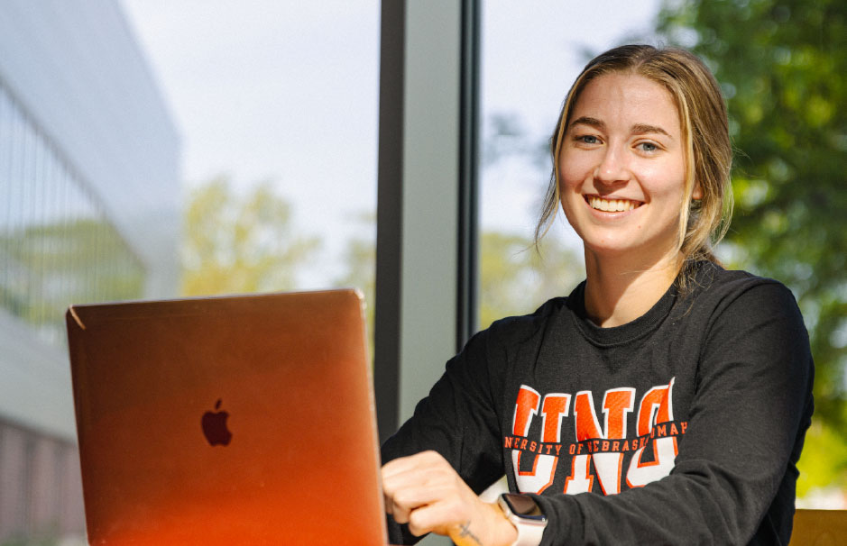 A smiling UNO student wearing a black sweatshirt works on a laptop near a window with trees and campus buildings in the background.