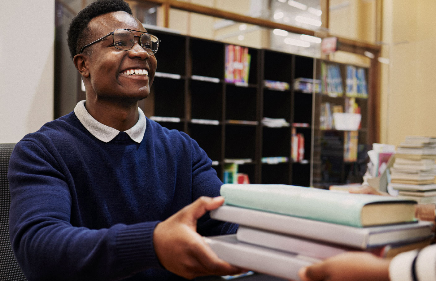Smiling library staff member handing a stack of books to a patron at a front desk, with shelves of books in the background.