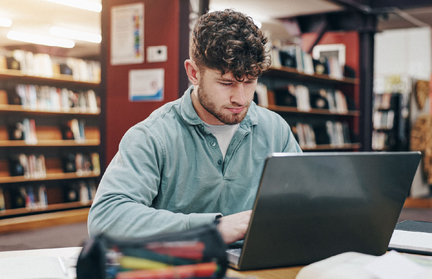 Student studying on a laptop in a library, focused on coursework, with bookshelves filled with academic materials in the background.