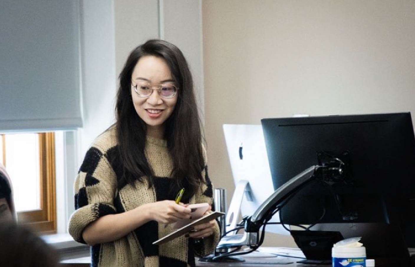 A woman wearing glasses and a beige and black patterned sweater stands in a classroom, smiling while holding a tablet and pen. She is positioned near a computer monitor and appears to be engaging with students off-frame.