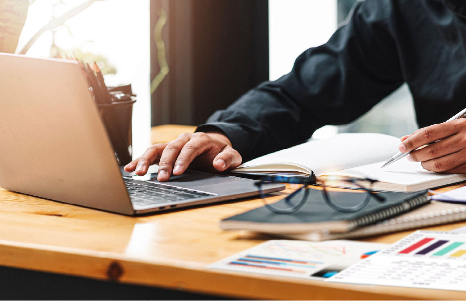 Close-up of a person using a laptop and taking notes at a desk, surrounded by charts, glasses, and a notebook.