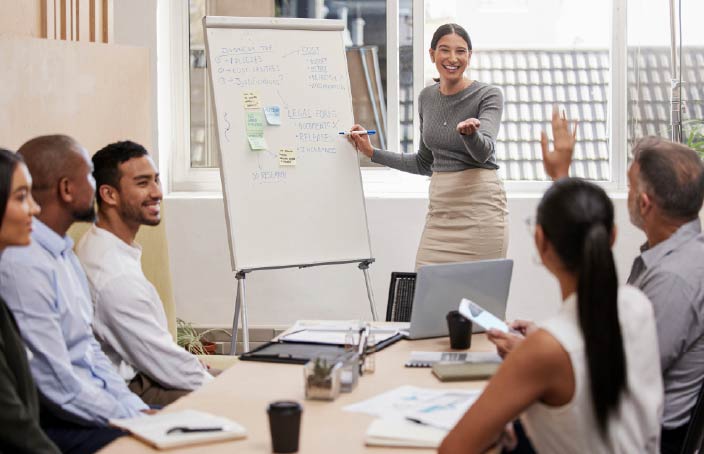 A woman stands smiling in front of a whiteboard during a presentation. She gestures to a group of diverse colleagues seated around a table. The whiteboard has sticky notes and handwritten ideas.