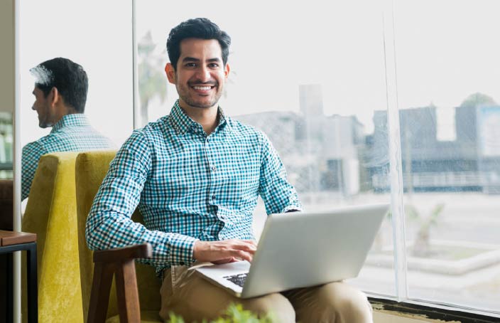 A smiling man in a blue plaid shirt sits in a modern chair by a large window, working on a laptop. He looks up from his work, with city buildings visible in the background.