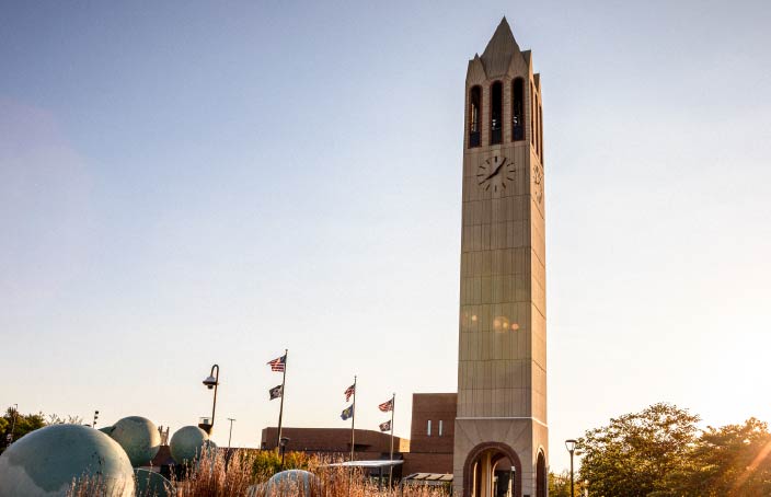 A tall clock tower stands on a university campus, with flags and trees around it. The sun is low in the sky, casting a golden light on the scene.