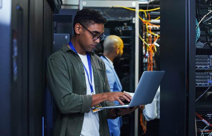A student works on a laptop in a server room surrounded by computer equipment, representing careers in information systems and technology management.