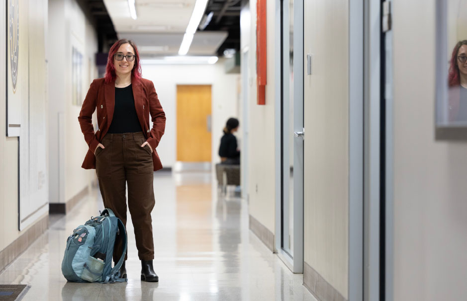 A student stands confidently in a campus hallway with a backpack at her feet, representing the flexibility and focus of the MPA curriculum at UNO.
