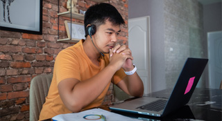 Asian man wearing headphones at kitchen table looking at laptop
