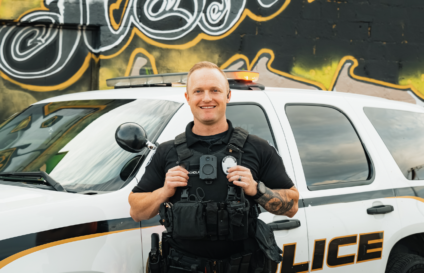 A smiling police officer wearing a black uniform and body camera stands confidently in front of a police vehicle. The background features a graffiti-covered wall, adding an urban feel to the scene.