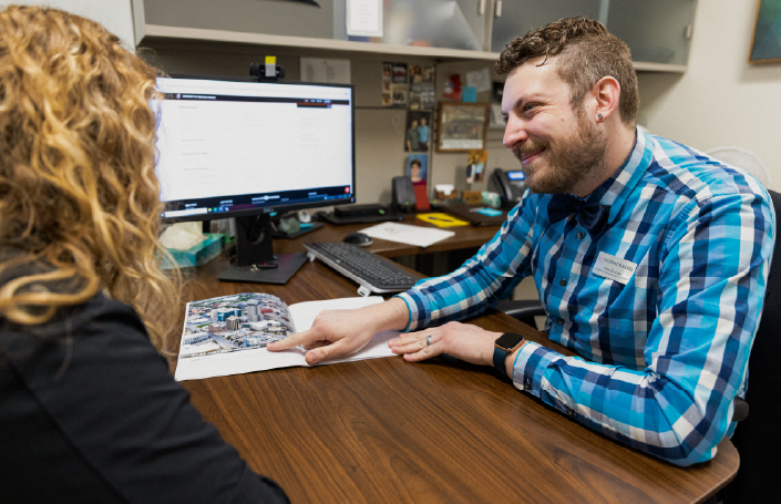 A friendly advisor in a blue plaid shirt engages with a student, pointing to a campus map in an advising session.