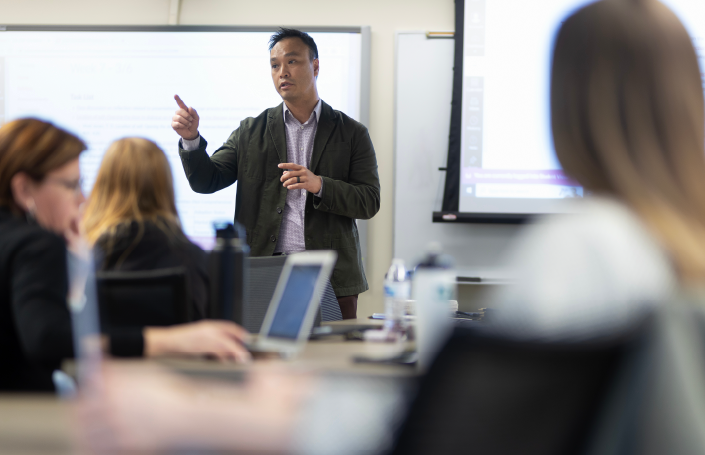 A faculty member leads a classroom discussion, gesturing as students with laptops listen attentively, representing engaged learning in higher education.