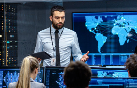 Man in a shirt and tie presenting in a security operations center, holding a tablet, with large monitors displaying world maps and digital network activity behind him.