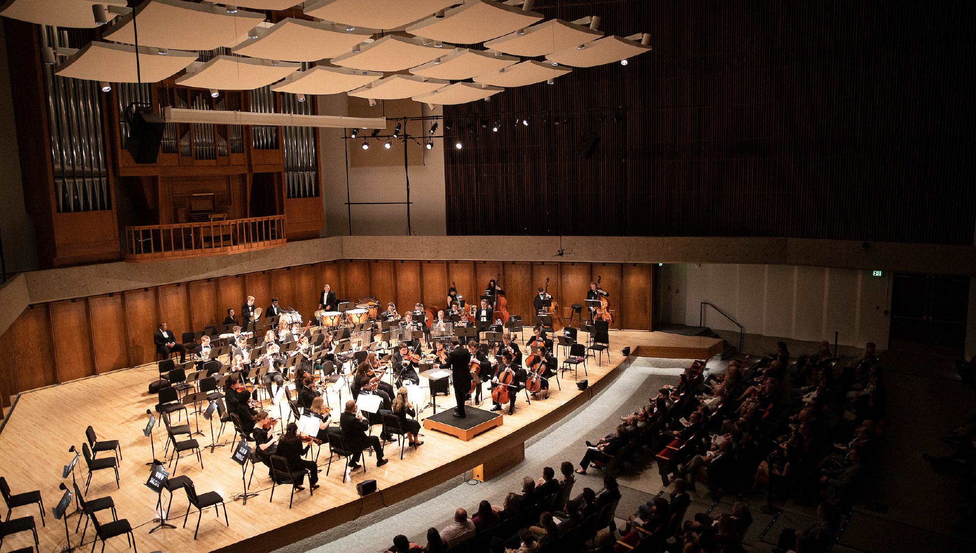 Orchestra performing on stage in the Strauss Performing Arts Center auditorium with audience seated in the hall.