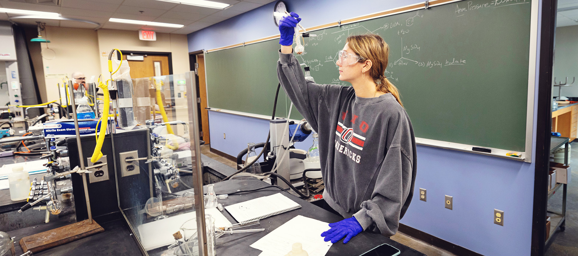 A student wearing safety goggles and gloves works with lab equipment in a chemistry classroom.