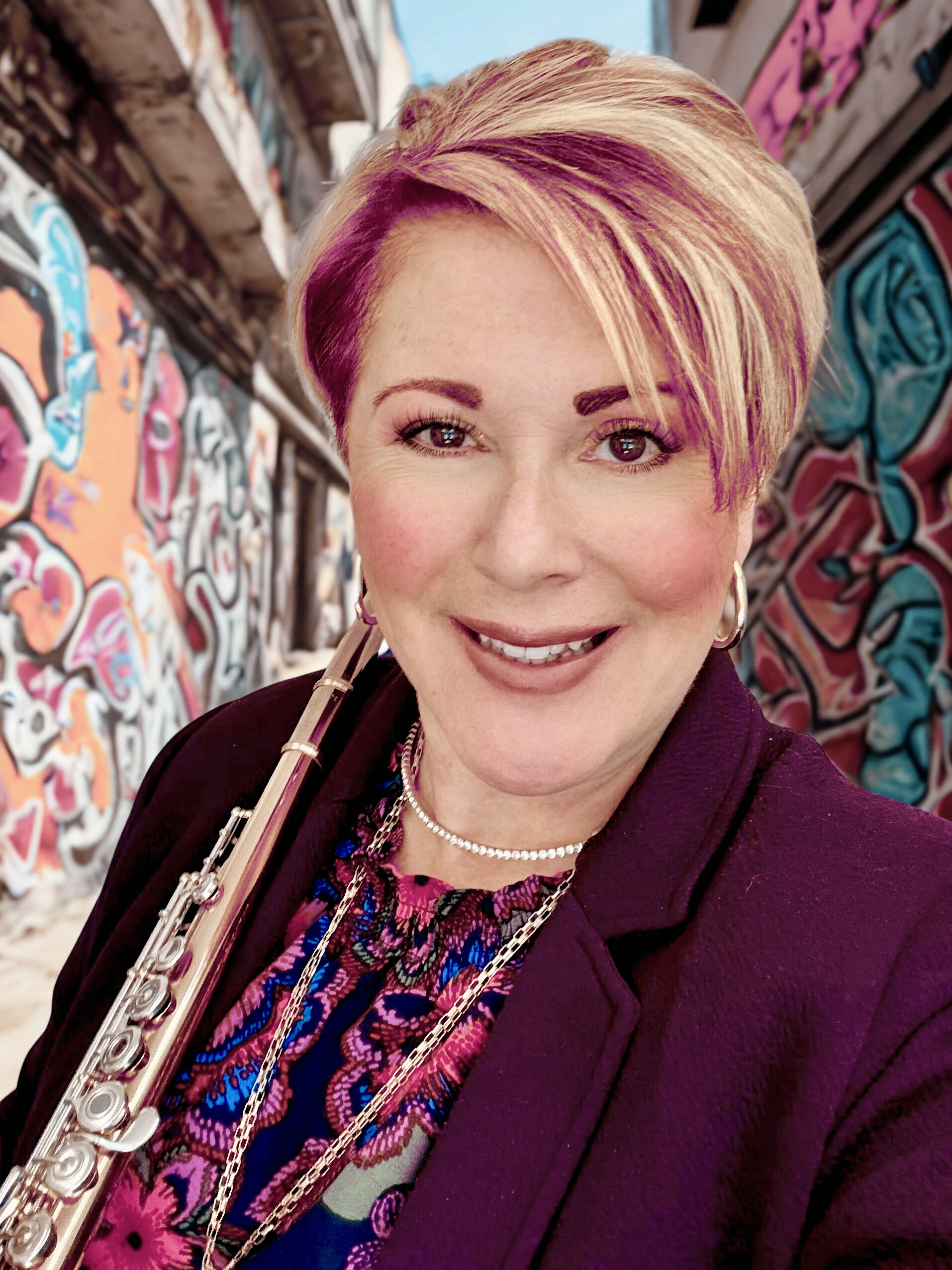 A woman with short, pink-highlighted hair holds a flute and smiles at the camera. She is wearing a purple blazer and a colorful patterned shirt. The background is a blurred image of a graffiti-covered alley.