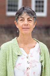 A smiling woman with gray hair, dangling earrings and a white patterned blouse with a green fuzzy cardigan.