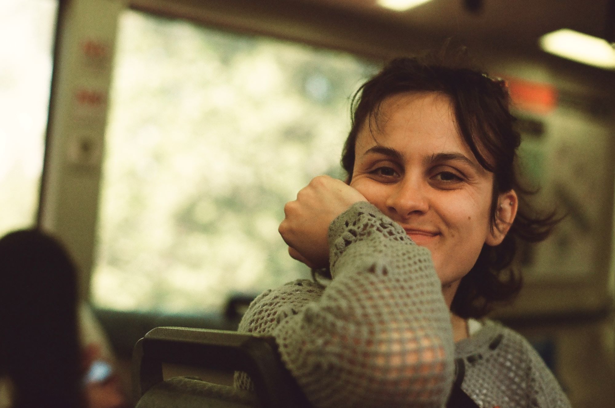 Chelsea is seen sitting in place of public transportation. Chelsea has short brown hair, with bangs and is wearing a long sleeve woven shirt. Chelsea's right arm rests on the top of her seat with her face resting in the palm of her right hand. There is a smile on Chelsea's face.