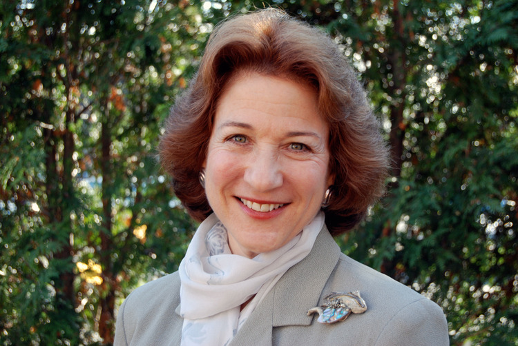 A shoulders-up shot of a light skinned woman with short brown feathered hair standing in front of a group of trees. She is wearing a white scarf, a grey suit jacket, and an iridescent broach.
